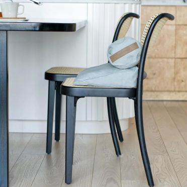 Poduszka klinowa i wałek w kolorze popiołu ułożone na krześle z plecionym oparciem przy kuchennym blacie.